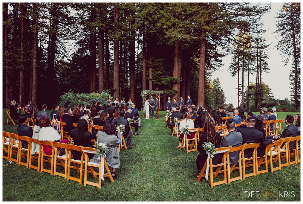 One image;: pullback image of bride and groom watched by guests standing in front of redwood trees.