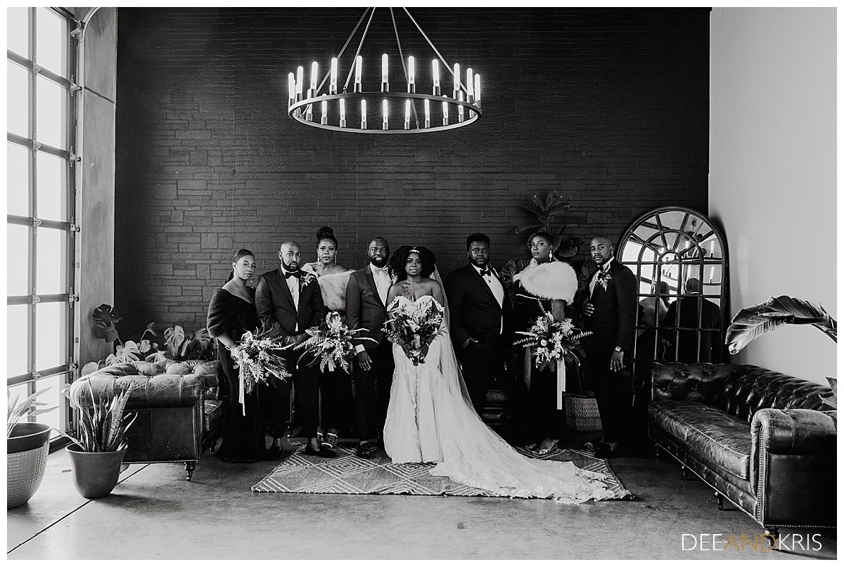 Single black and white image of wedding party standing in lounge seating area with large window, chandelier, brick wall, arched mirror and couch seating.