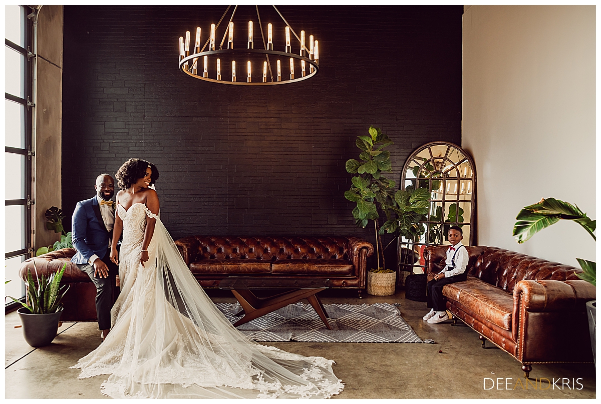 Single image of bride and groom in lounge area with large window, brick wall, chandelier, mirror and couch seating with son looking on.