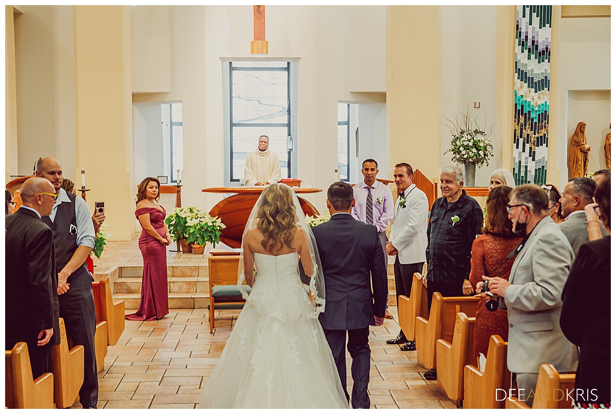 Single POV image from beginning of aisle looking toward front of aisle at bride and father from behind walking toward groom as he watches.