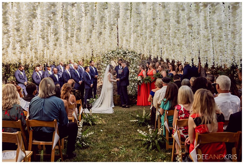 One image of pride and groom at ceremony under curtain of wisteria holding hands for Tie The Knot: Dream Wedding Guide blog post.