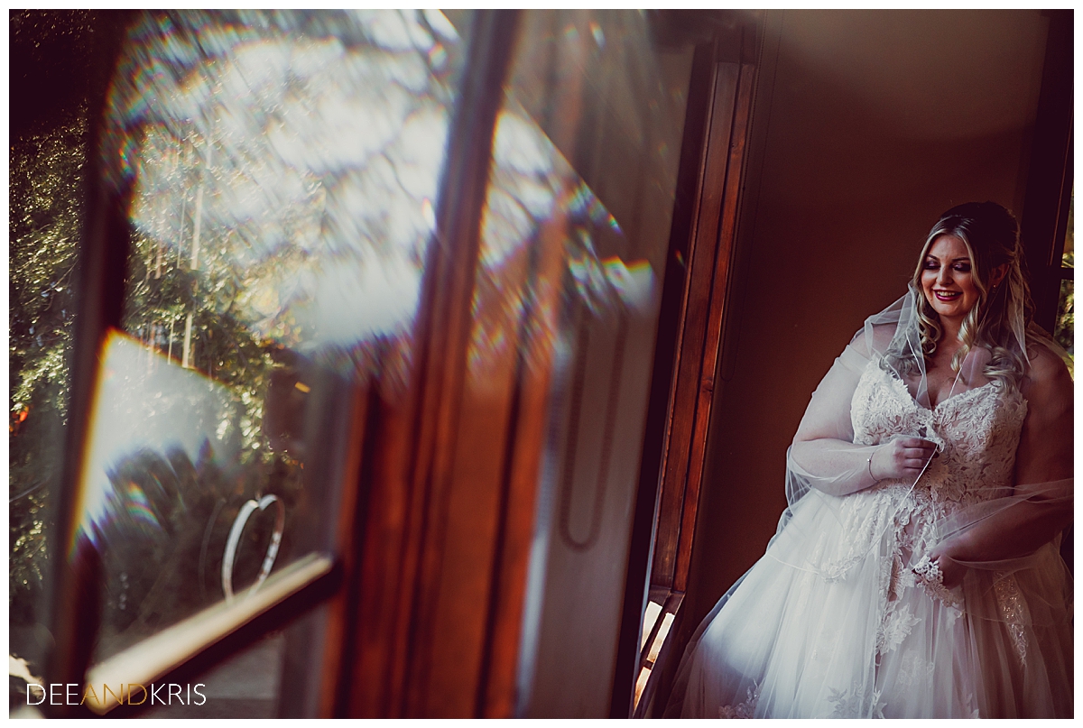 Single image of bride standing in window looking down and smiling.